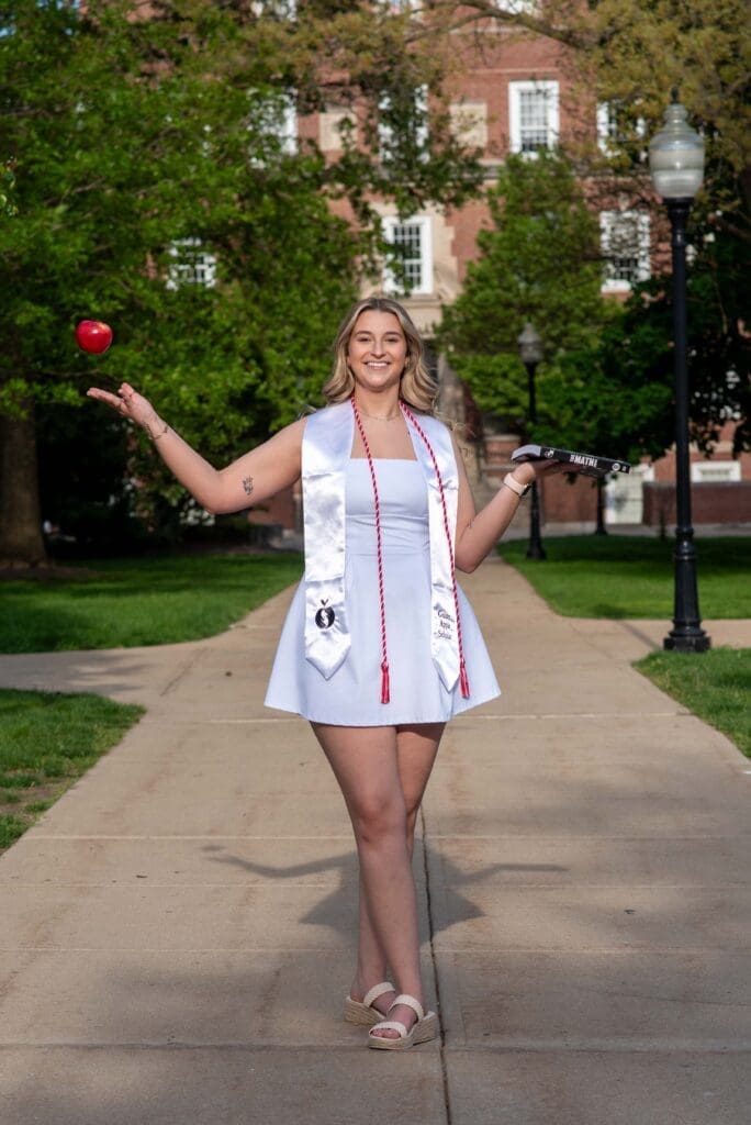 college graduate in a white dress with a book in hand a tossing an apple